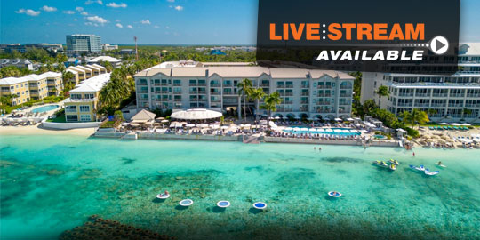 Panoramic view of the Grand Cayman Marriott Resort, a beachfront resort on Grand Cayman Island. The resort features a multi-story building with balconies overlooking a swimming pool complex, white sand beach, and the turquoise Caribbean Sea.