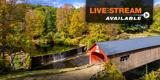 A historic covered bridge spans a cascading waterfall amidst vibrant fall foliage in Manchester, Vermont, the picturesque setting for CME/CE conferences with live stream options.