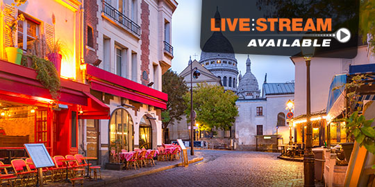 Tranquil Parisian street scene with cafes and shops lining a narrow cobblestone road, with the Sacré-Coeur Basilica visible on a hill in the background.