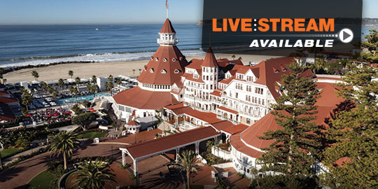 Exterior view of the historic Hotel del Coronado, a beachfront resort in San Diego, California, with a red tile roof, Victorian architecture, and trees lining the entrance.