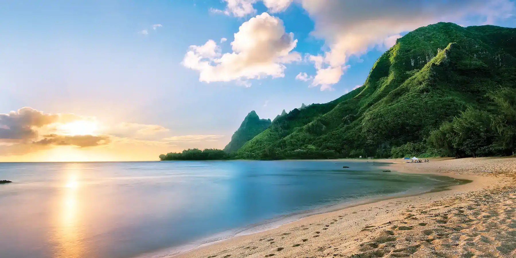 A tranquil Hawaiian beach with golden sand, lush green mountains in the background, and a calm ocean reflecting a colorful sunset.